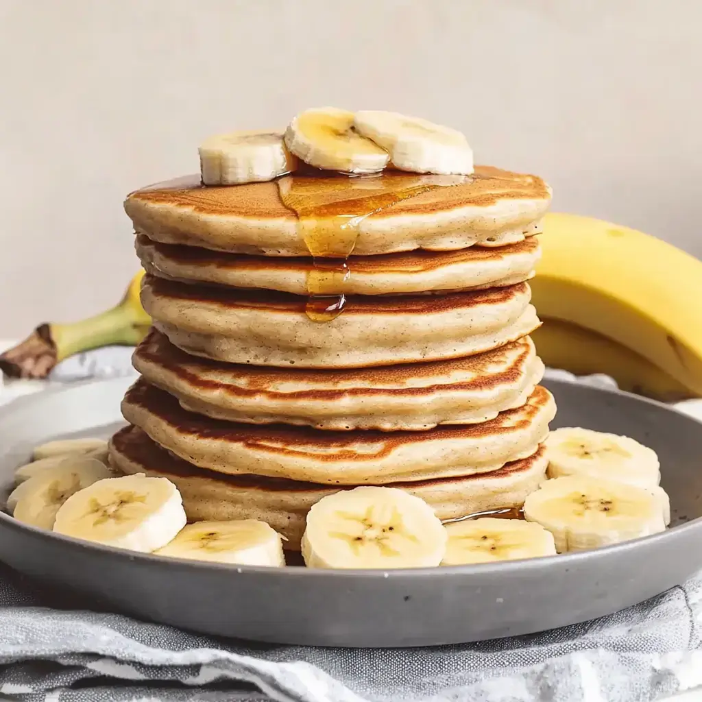 A large stack of pancakes on a plate, topped with banana slices and syrup, next to a banana.