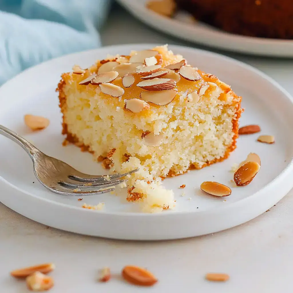 A slice of almond-topped cake on a white plate, sprinkled with almond flakes, sitting by a fork.