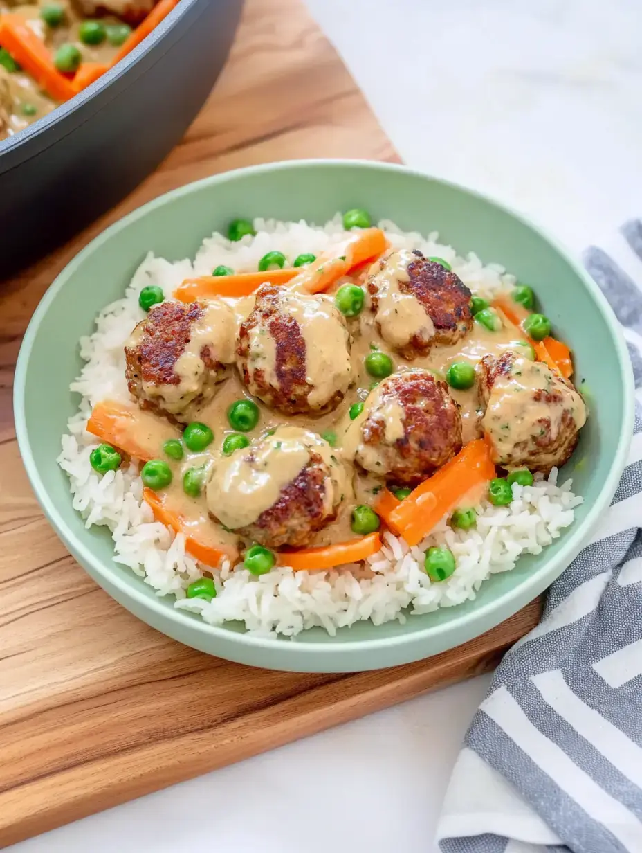 A plate of rice and meatballs with creamy sauce, peas, and carrot strips in a green bowl.