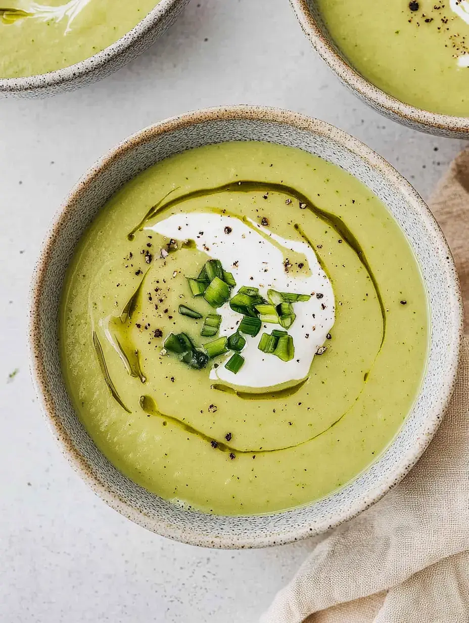 A bowl of creamy green soup topped with sour cream, chopped chives, and a drizzle of olive oil.